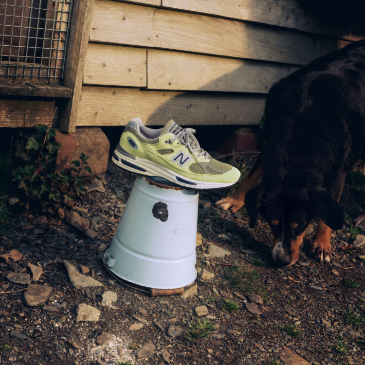 A New Balance Made in UK sneaker sitting on an upturned white bucket in a rustic farm environment next to a dog with dirt and leaves on the ground and an old shed with weatherboard cladding behind. 