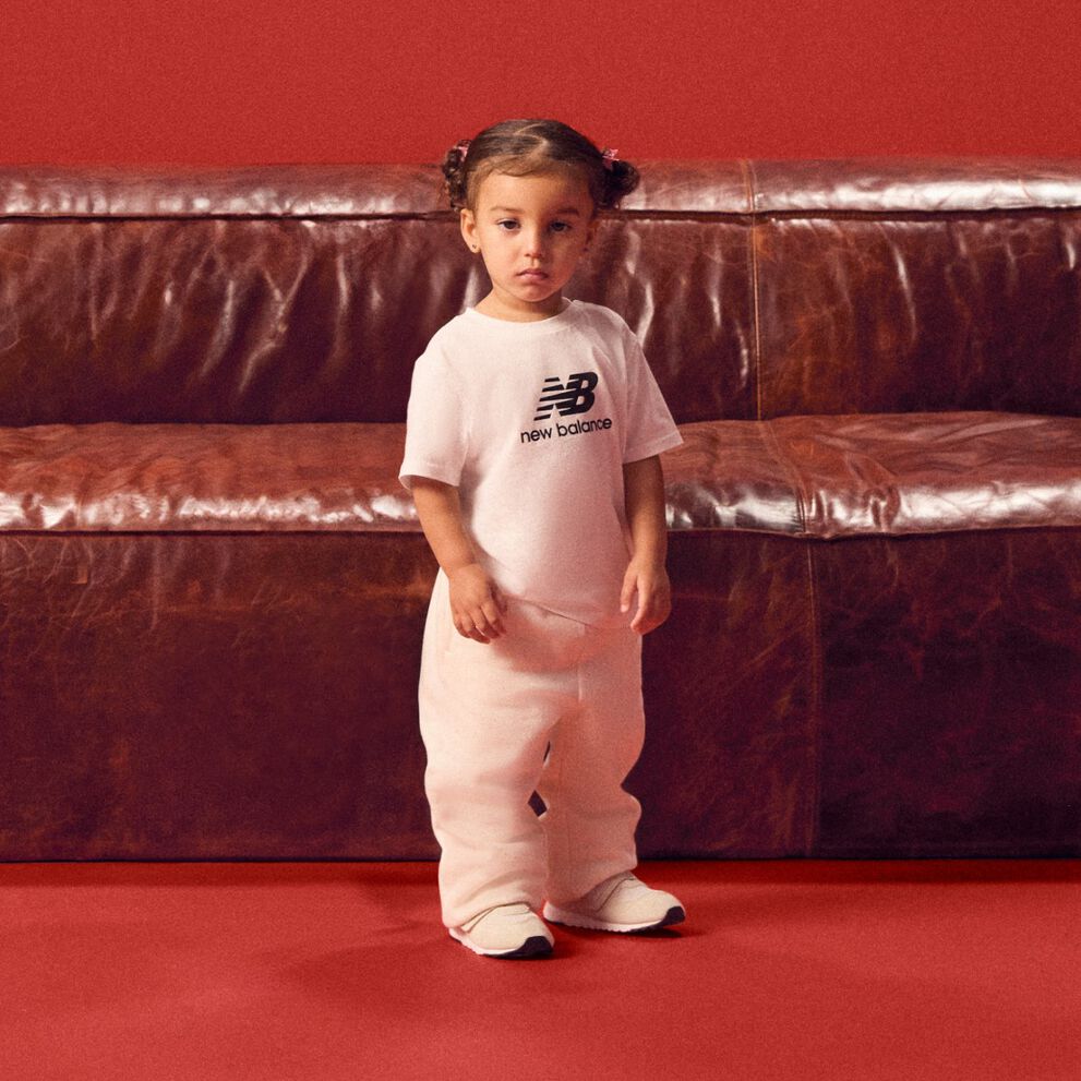 Baby standing in front of a red couch wearing a white tshirt.