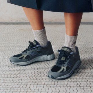 Feet view of a woman wearing a pair of New Balance Made in UK sneakers standing on a white tiled floor.