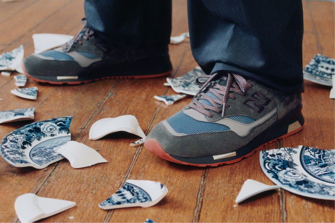 Knee down view of a man wearing a pair of New Balance Made in UK sneakers standing on a rustic wooden floor with smashed blue and white ceramic crockery all over the ground and white cabinets in the background. 